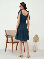 Woman wearing a dark blue dress with bow straps, standing in a minimalistic room.