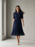 Woman wearing a blue dress standing in front of a white wall with a window.