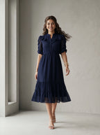 Woman wearing a blue dress standing in front of a white wall with a window.
