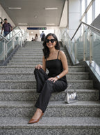 A woman wearing a black camisole top and black wide-leg trousers sits on a wide staircase in a modern indoor transit station. She is wearing brown pointed-toe heels and a silver handbag rests beside her.